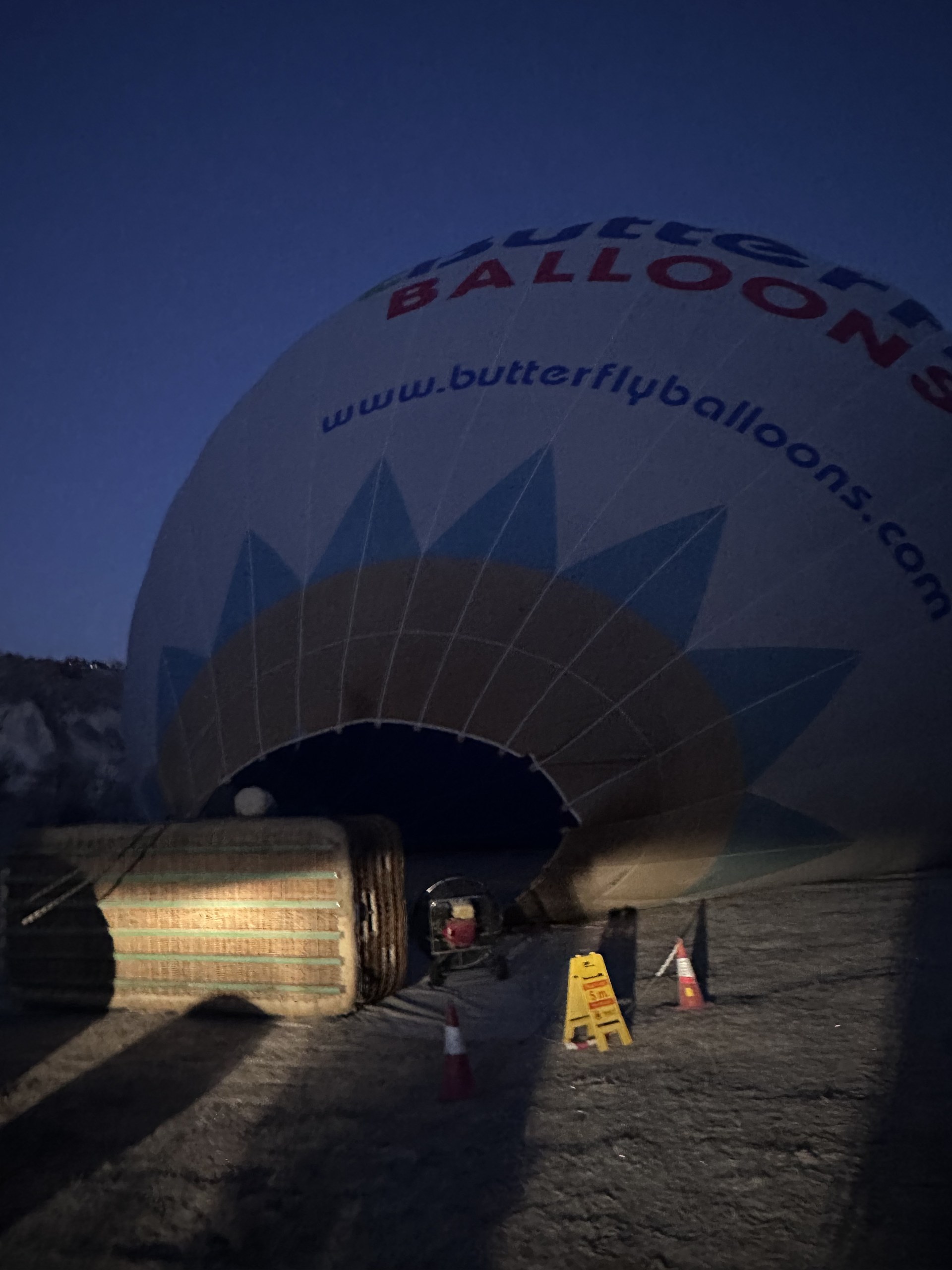夜明け前、Butterfly Balloons の球皮が地面に広がり、膨らみ始める。バスケットはまだ横倒しで、球皮には butterflyballoons.com の文字。