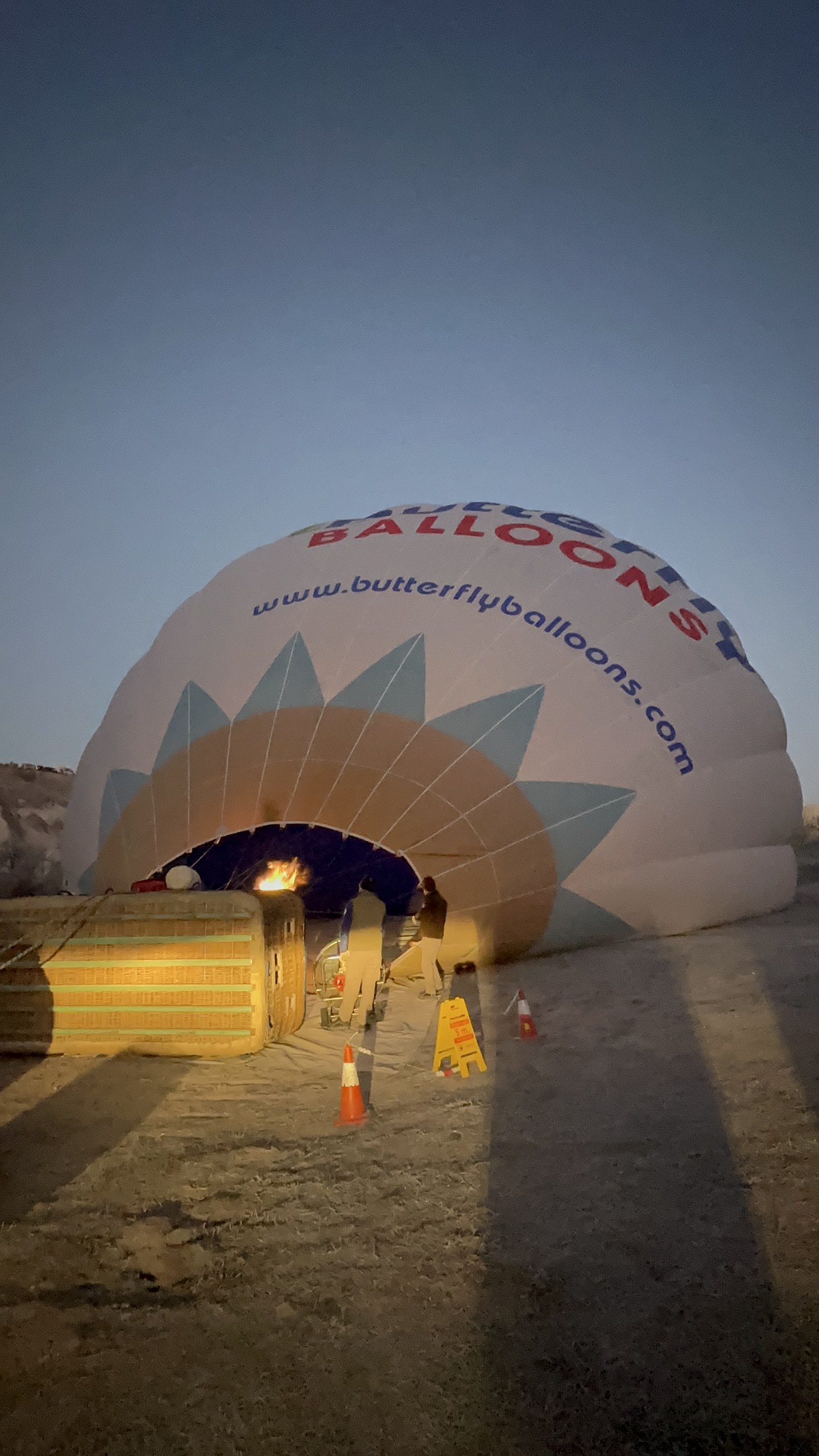 隣の Butterfly の気球も充気加熱中：バーナーが球皮の口に炎を吹き込み、球皮はもう大半が膨らんだ。バスケットの脇にはコーンと案内板。