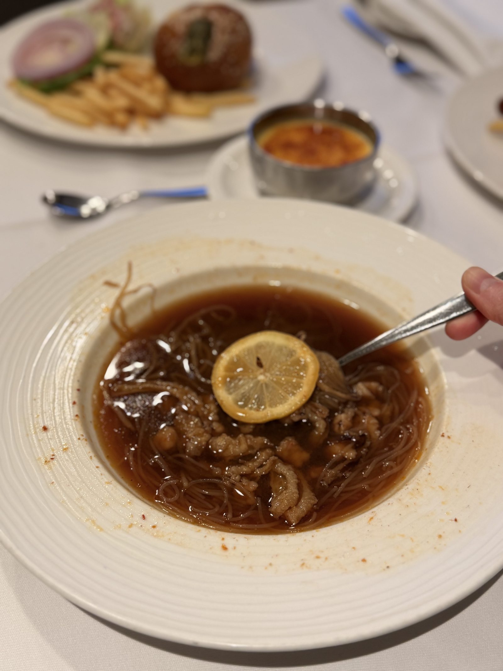 麺のスープのアップ。濃い色のスープに細い春雨と肉のスライス、レモンの輪切りが浮かびます。