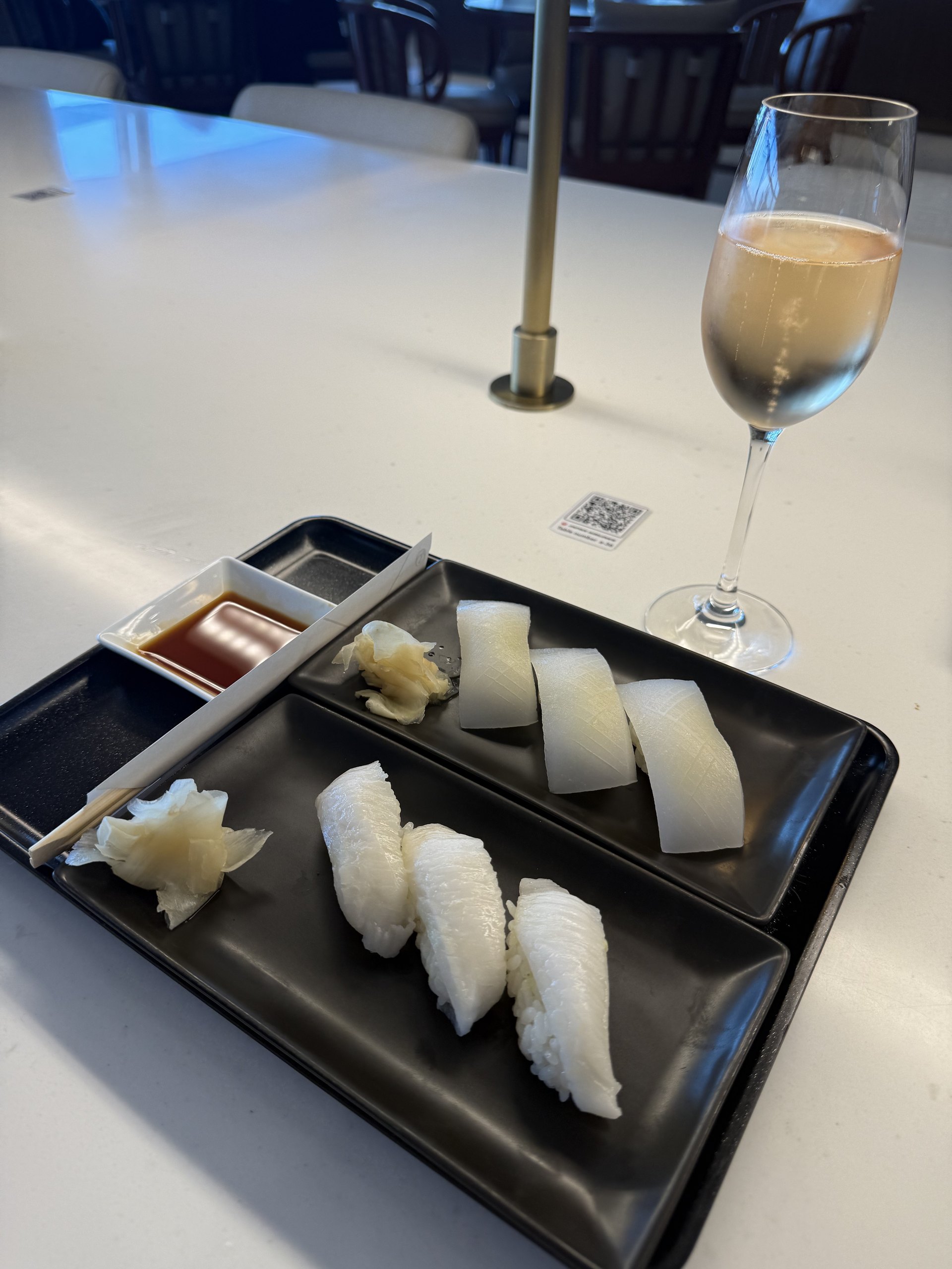 Two plates at once on the counter: three cuttlefish, three white fish, a glass of champagne beside them, and the ordering QR code on the white counter.