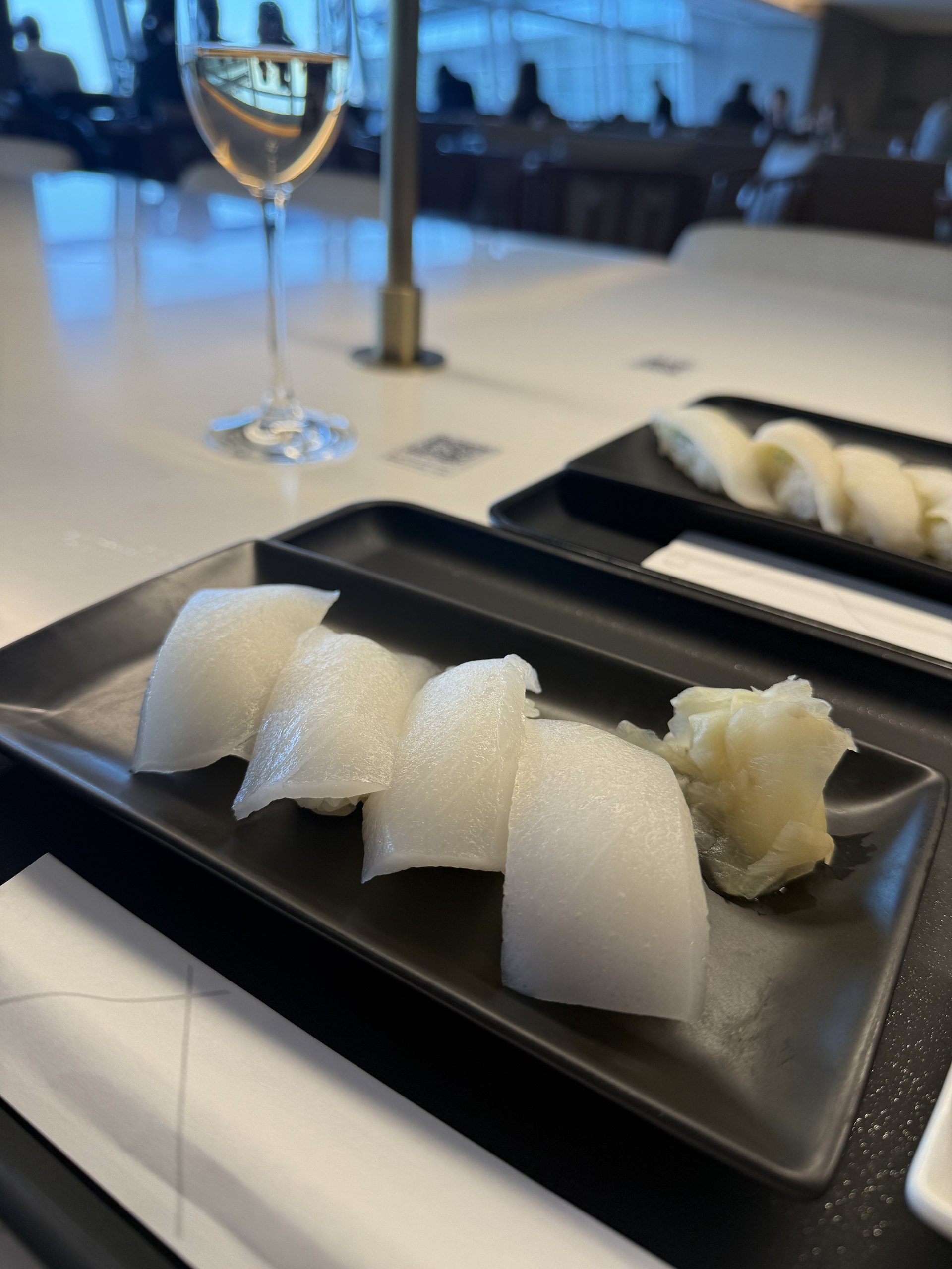 Cuttlefish, four pieces, up close: the translucent flesh scored, ginger alongside, the hall and a glass of white blurred behind.