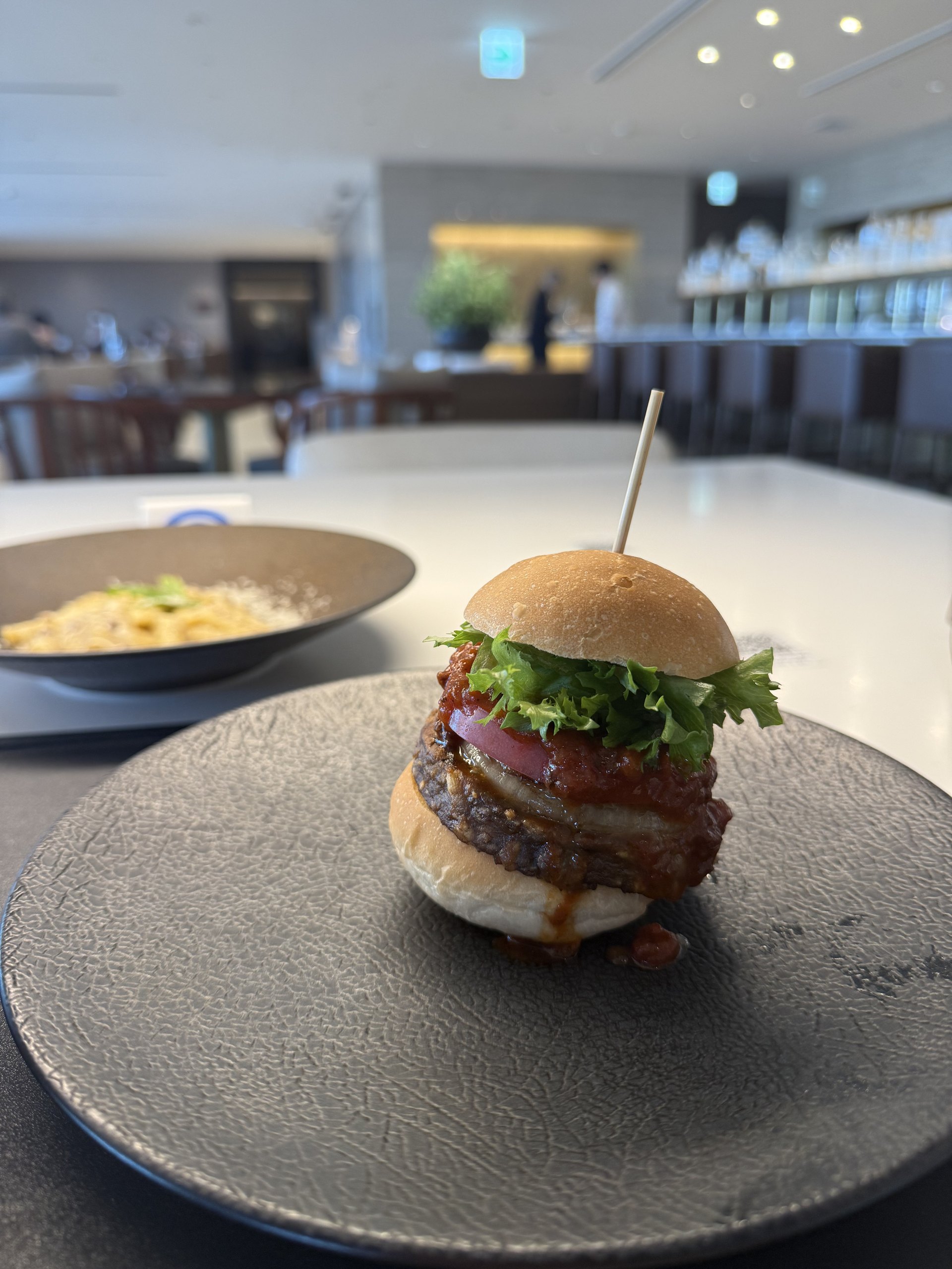 The burger, stacked high — beef patty, tomato, lettuce and sauce held by a skewer — with a plate of pasta beside it.