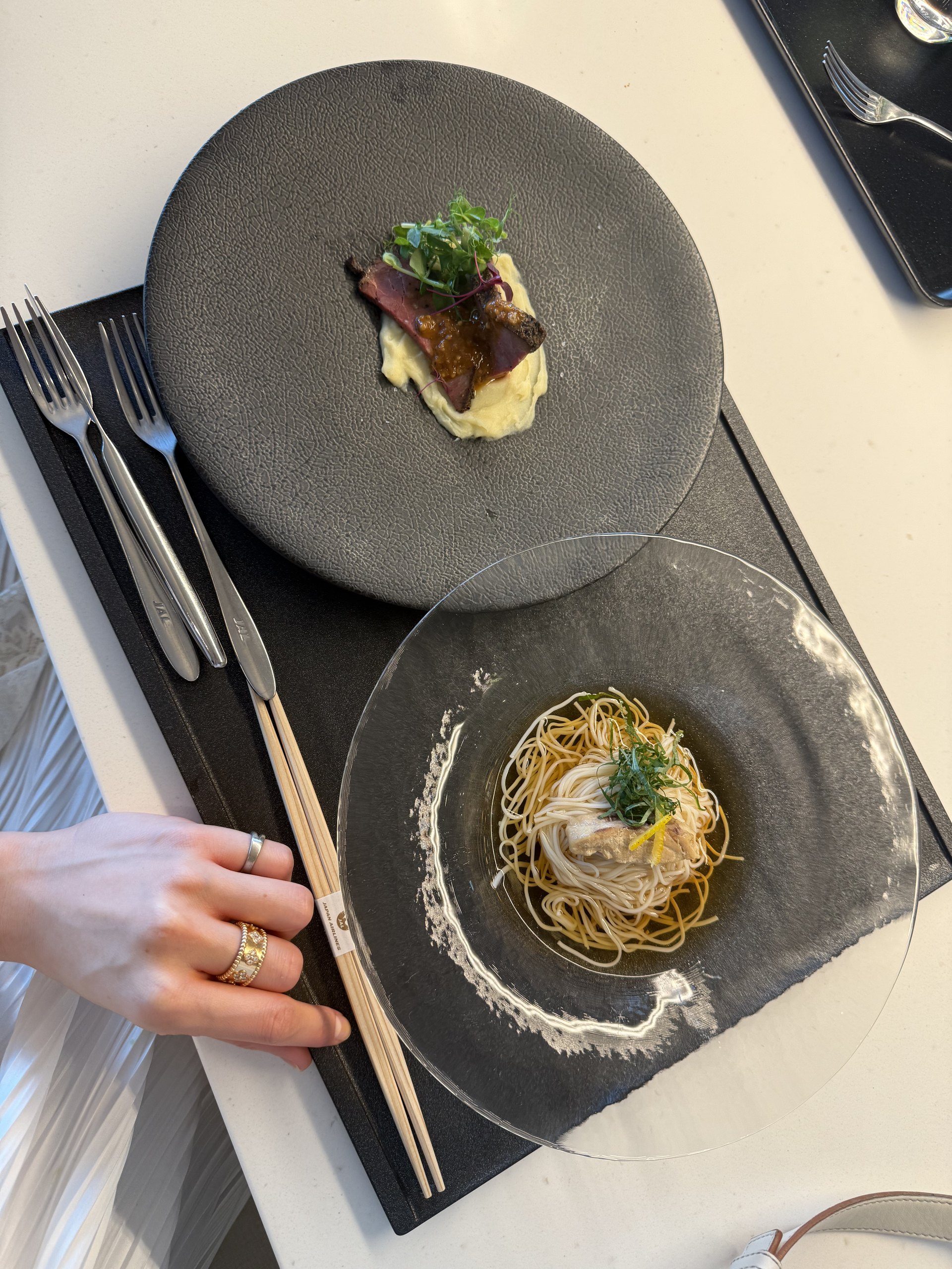 Two dishes together: roast beef slices on mashed potato with sprouts, and chilled fine noodles in a glass plate topped with a slice of fish, shiso and lemon zest. Even the forks and chopstick band carry JAL's name.