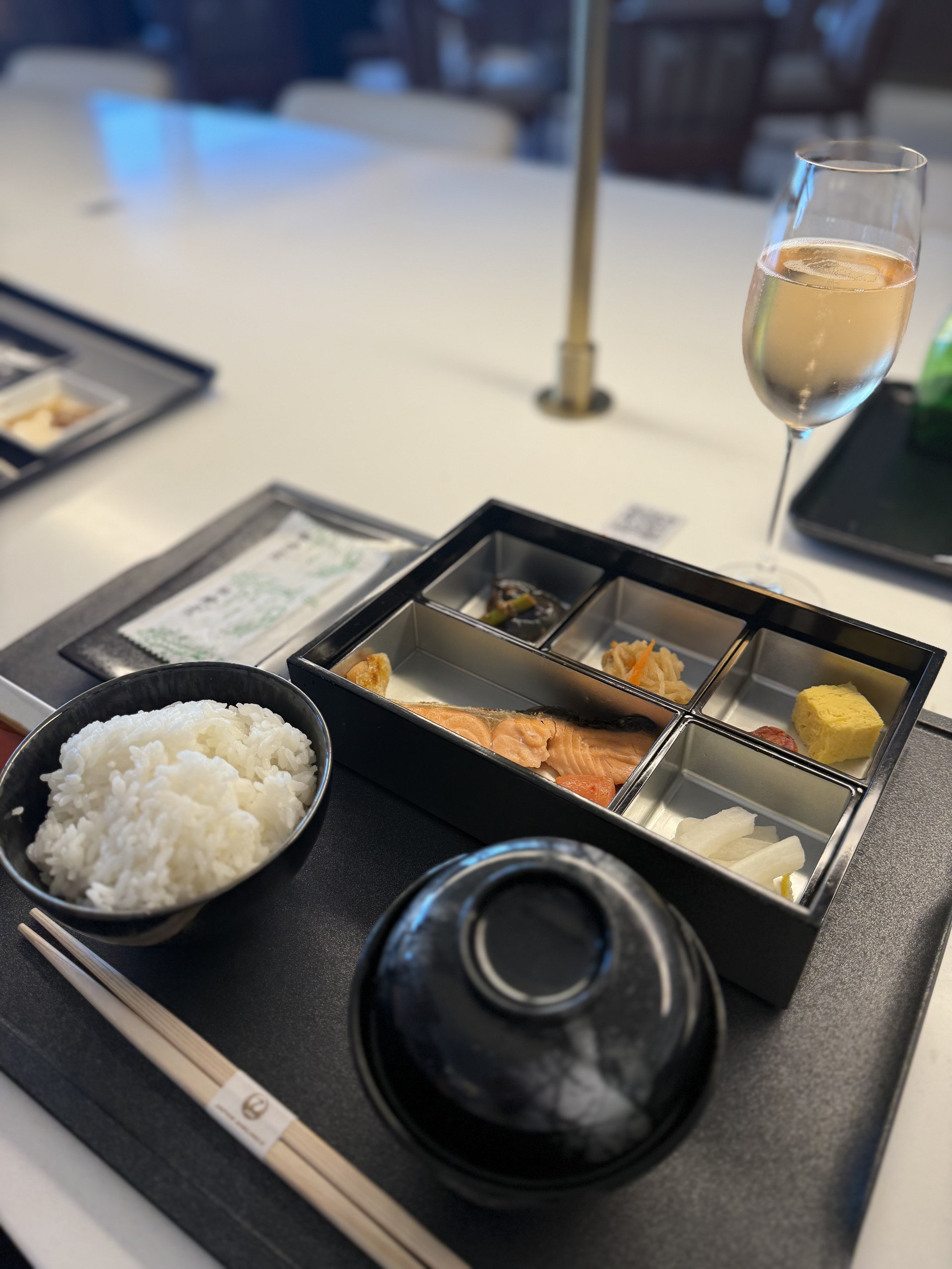A Japanese bento: grilled salmon, tamago, pickles and simmered vegetables in their compartments, with a bowl of rice, a lidded soup bowl, and champagne alongside.